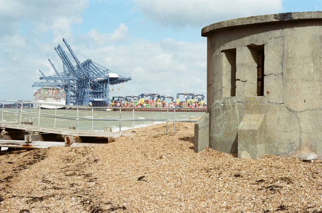 A crumbling Second World War concrete pillbox on the shingle beach at Felixstowe, with container ships and port cranes visible across the water