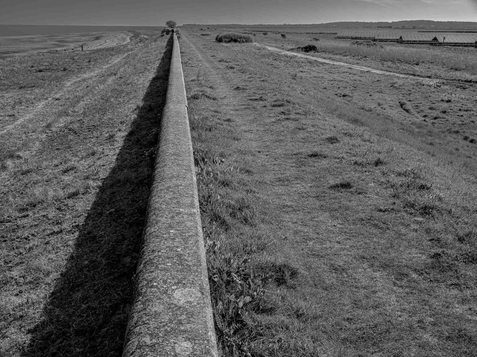 Black and white photograph of a concrete sea wall dividing coastal marshland from Cleve Hill Solar Park, Kent, with the Swale estuary visible on the left horizon