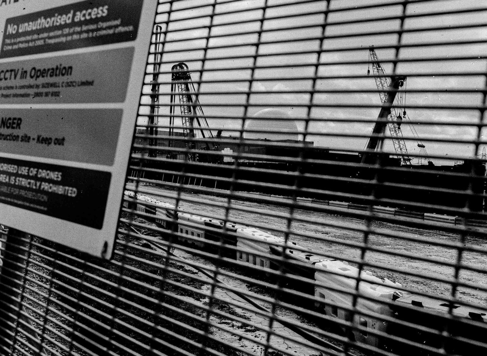 The Sizewell C construction site viewed through a security fence, with warning signs visible and construction cranes and cleared earth behind the mesh