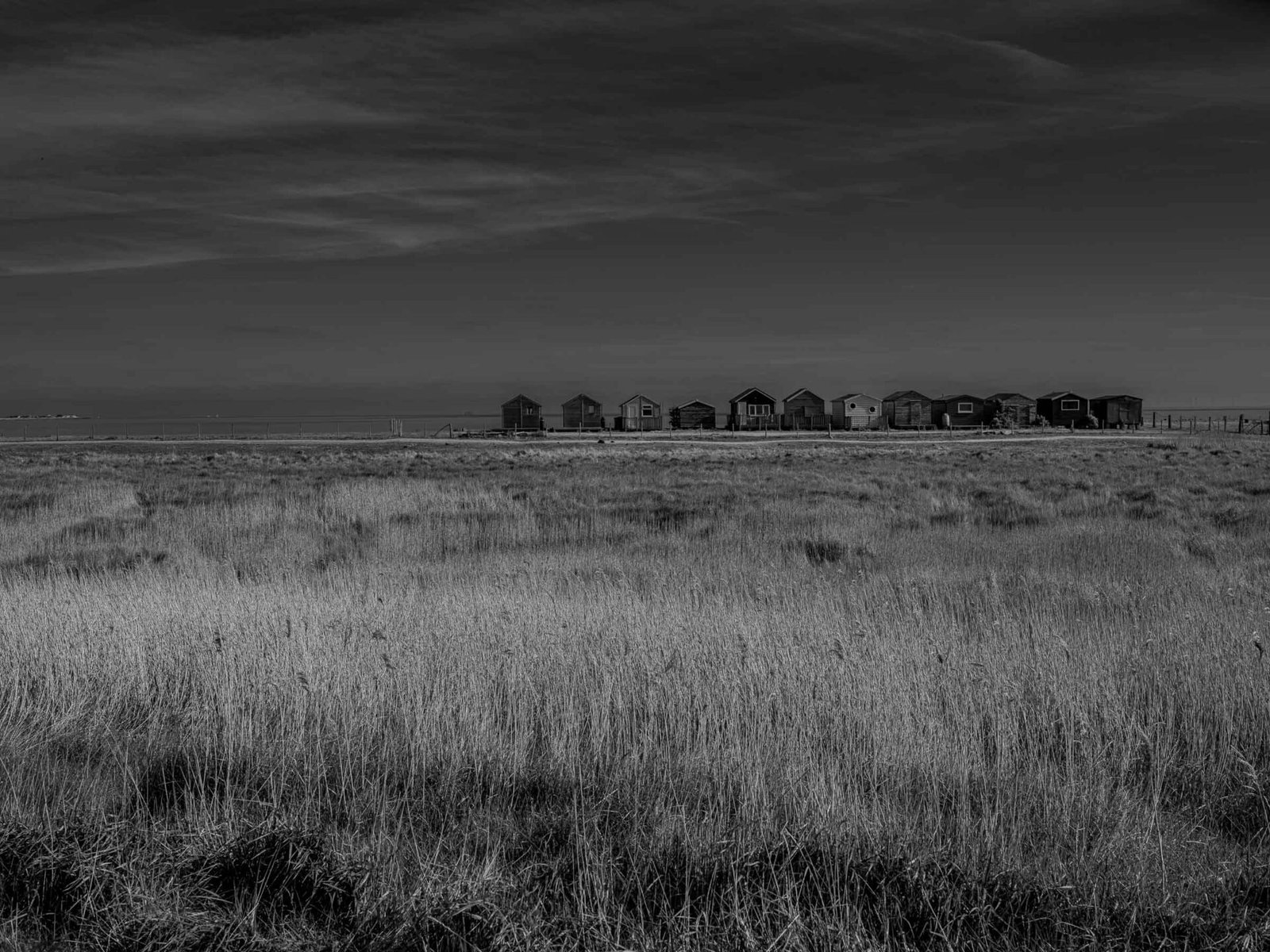 Black and white photograph of beach huts on the Swale estuary shoreline, Kent, with dark dramatic sky above coastal marshland