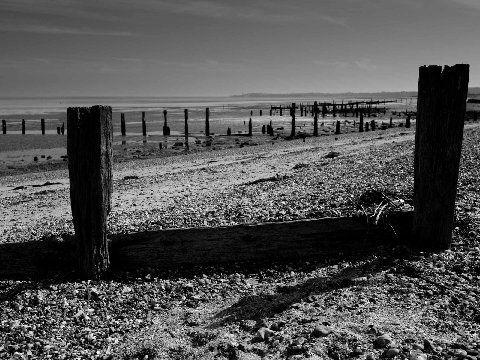 Black and white photograph of decaying wooden groynes on the Swale estuary at low tide, Whitstable visible on the distant horizon