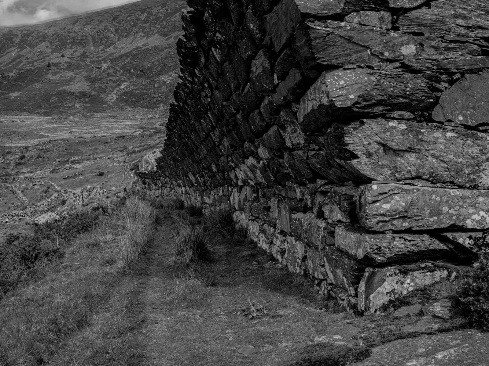 A weathered stone retaining wall holds back the slate waste at Gorseddau Quarry, with the valley falling away beyond it.