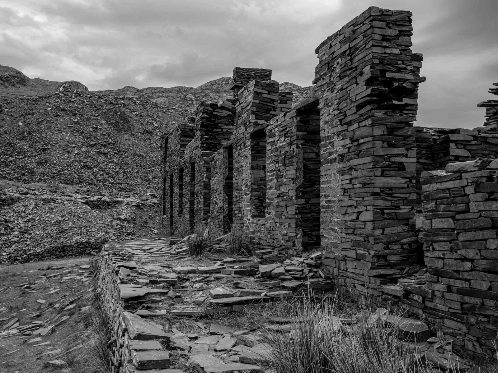 A row of roofless slate-built compartments at Cwmorthin Quarry, receding into the distance with a mountain behind.