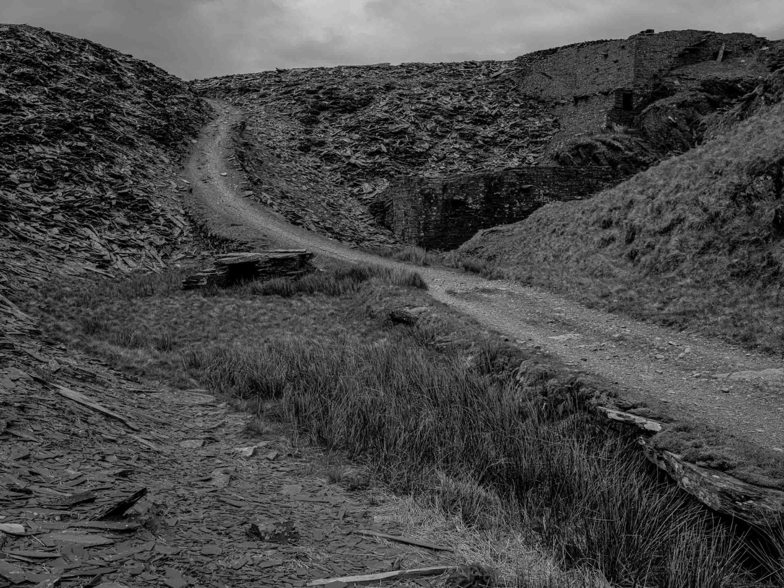 A path cutting through mounds of broken slate waste toward a ruined building at Conglog Quarry.