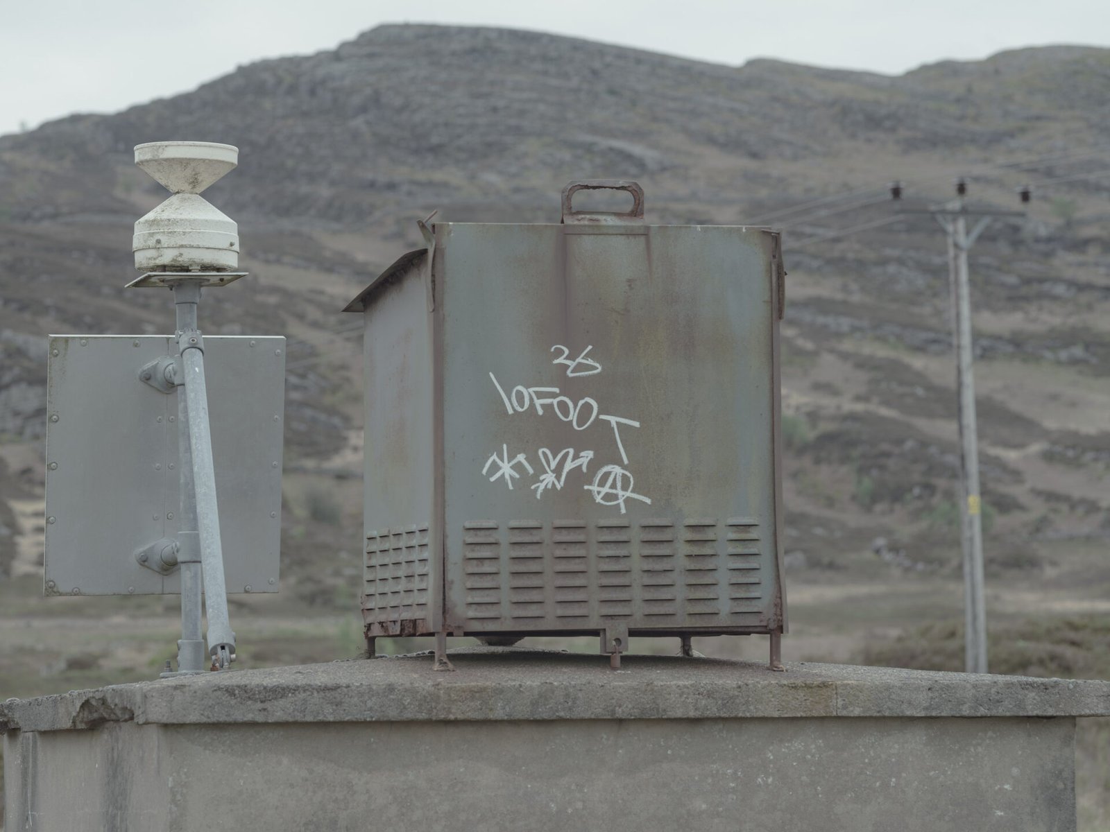 Rusted nuclear monitoring equipment with graffiti on concrete plinth, bare mountain behind, Trawsfynydd