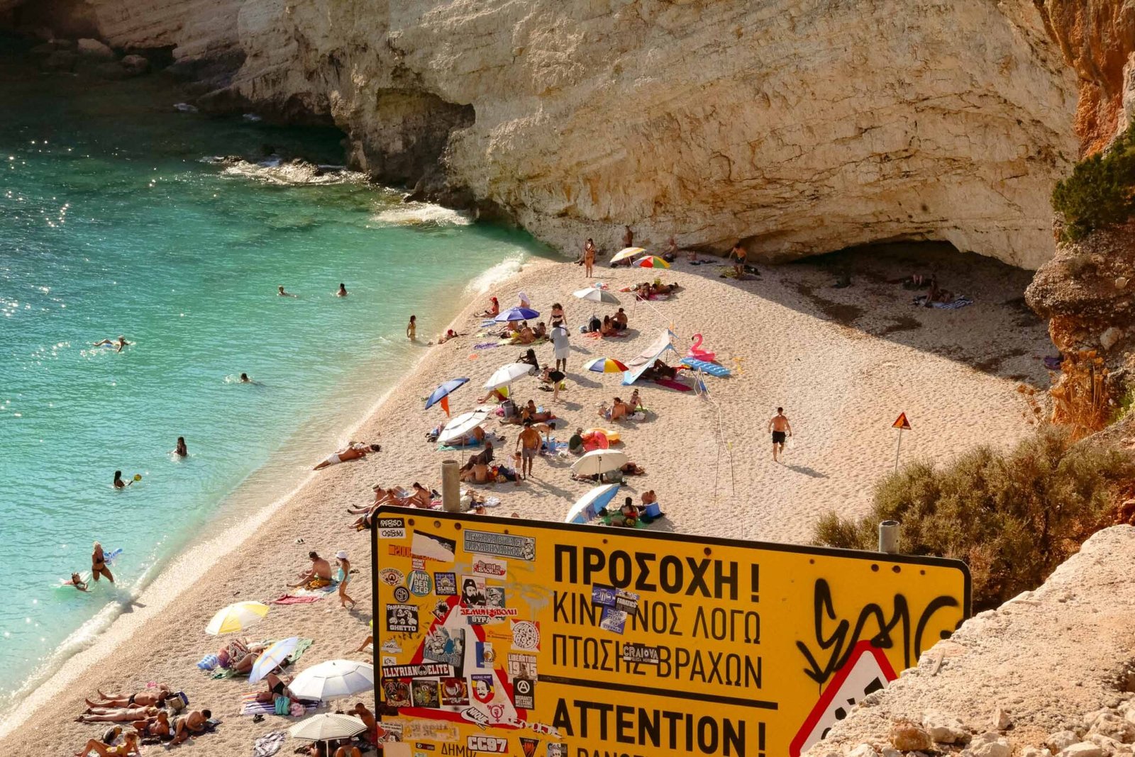 Sticker-covered rockfall warning sign overlooking a crowded turquoise beach beneath cliffs, Lefkada, Greece