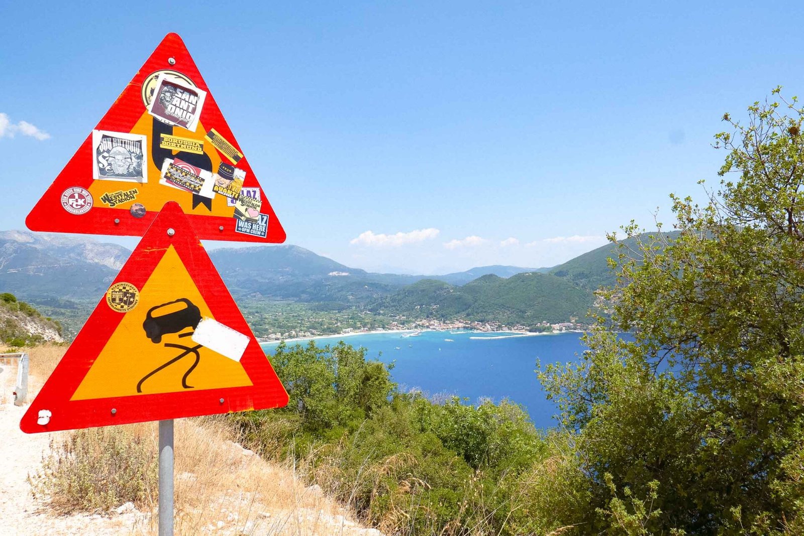 Road warning signs covered in football fan stickers overlooking a bay on Lefkada, Greece