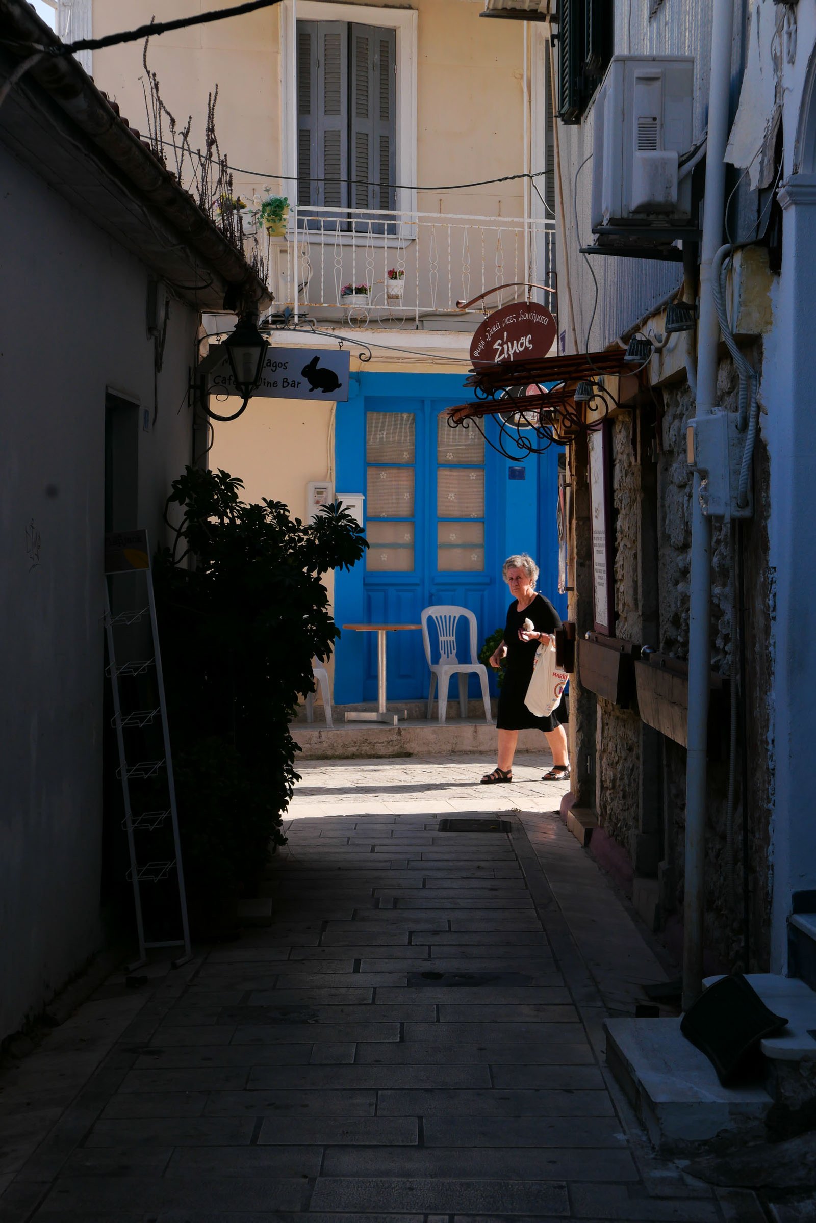 Woman walking past a blue-shuttered doorway in a sunlit alley in Lefkada Town, Greece