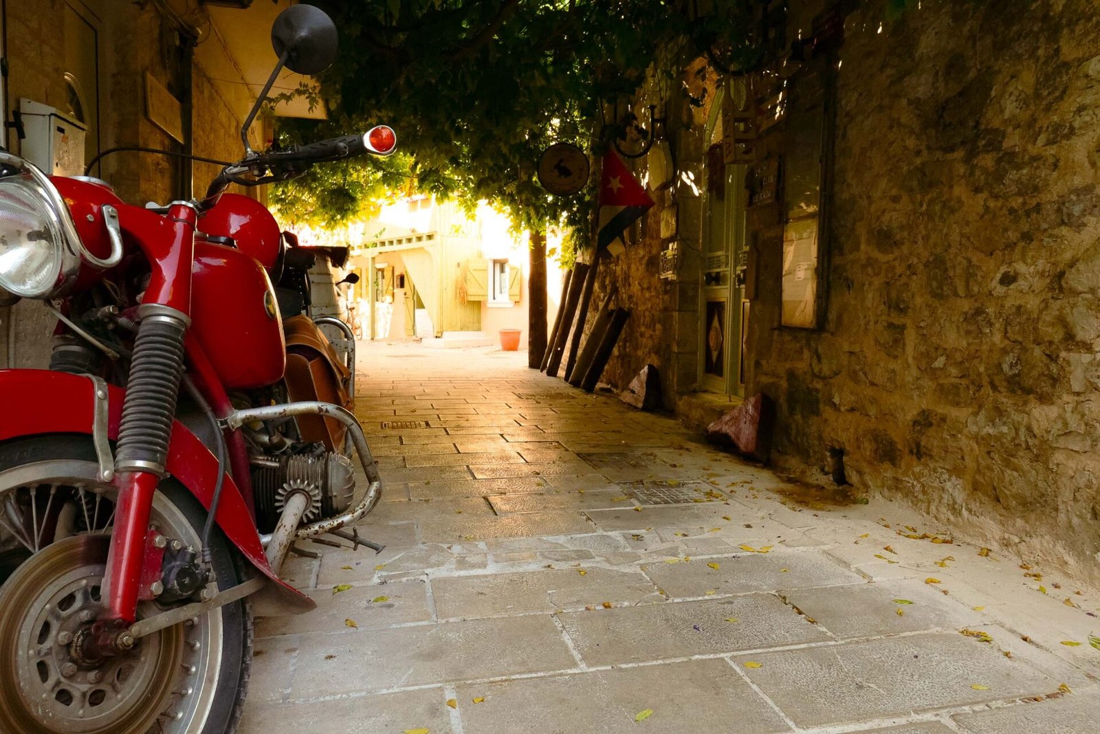 Red vintage motorbike parked in a shaded stone alleyway leading to sunlit Lefkada Town, Greece