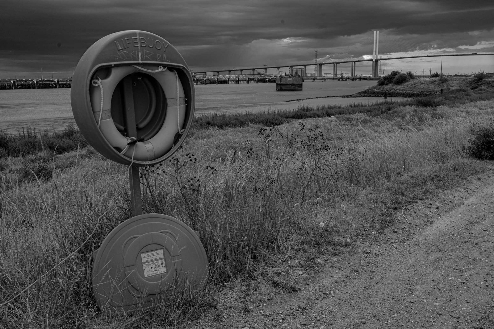 Thames waterside with lifebuoy mounted on weathered post in foreground; Dartford Crossing bridge visible across grey water under dramatic clouded sky; saltmarsh vegetation.