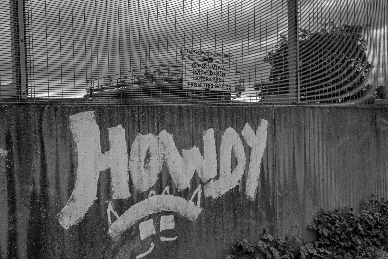 Lower half of industrial corrugated metal wall covered in large white "HOWDY" graffiti; upper half shows Thames Water Authority warning signage about sewer outfall; industrial mesh fencing visible.