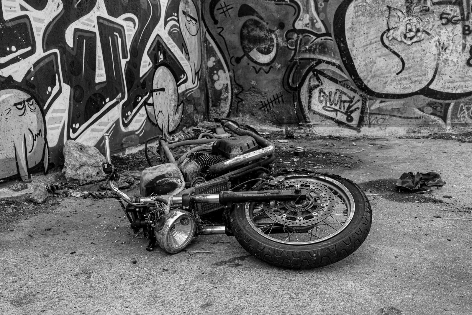 Charred, burnt-out motorcycle frame and engine components lying abandoned among debris; graffiti-covered industrial walls behind; minimal remaining structure, skeletal metal remains.