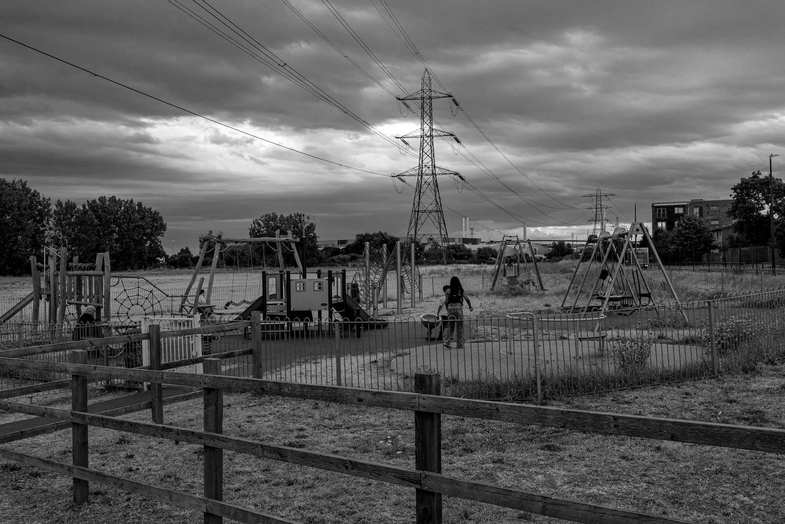 Children's playground with equipment in foreground; single child visible on equipment; massive electrical transmission pylon dominating background; Dartford Crossing bridge visible on horizon; open field setting; dramatic clouded sky.