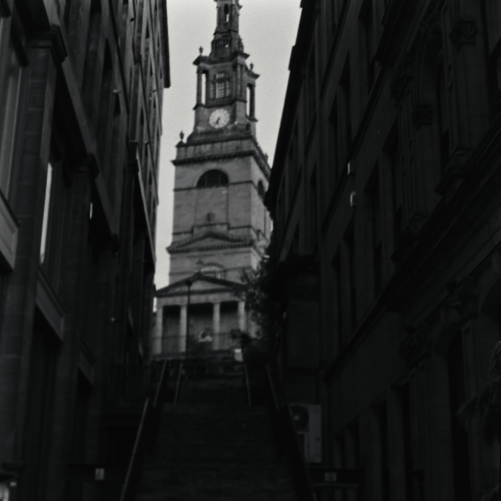 Black and white film photograph of All Saints Church tower in Newcastle, framed between dark buildings at the top of a narrow stone stairway