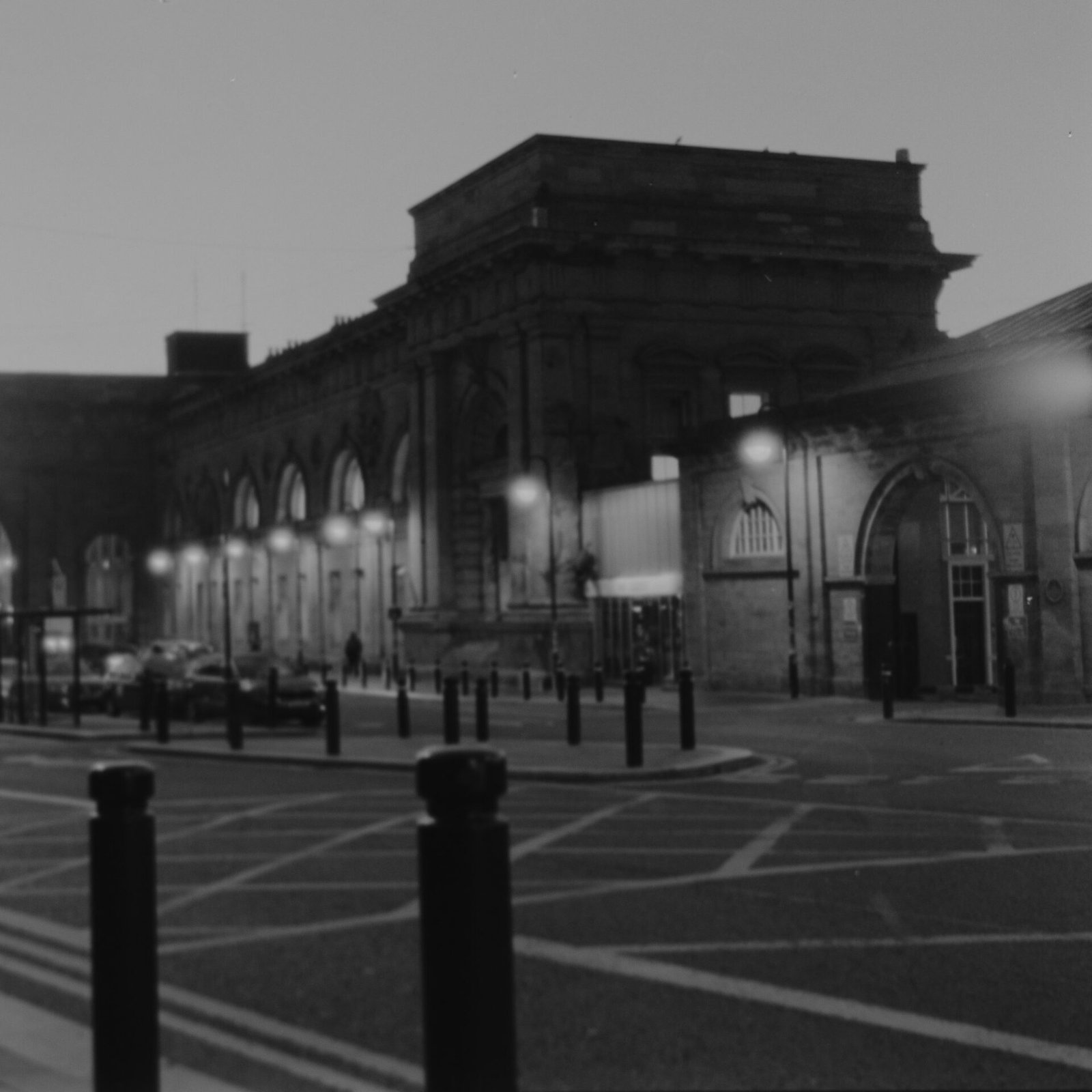 Newcastle Central Station portico before sunrise on black and white film, street lamps glowing along the arcade, a lone figure walking past and bollards in the foreground