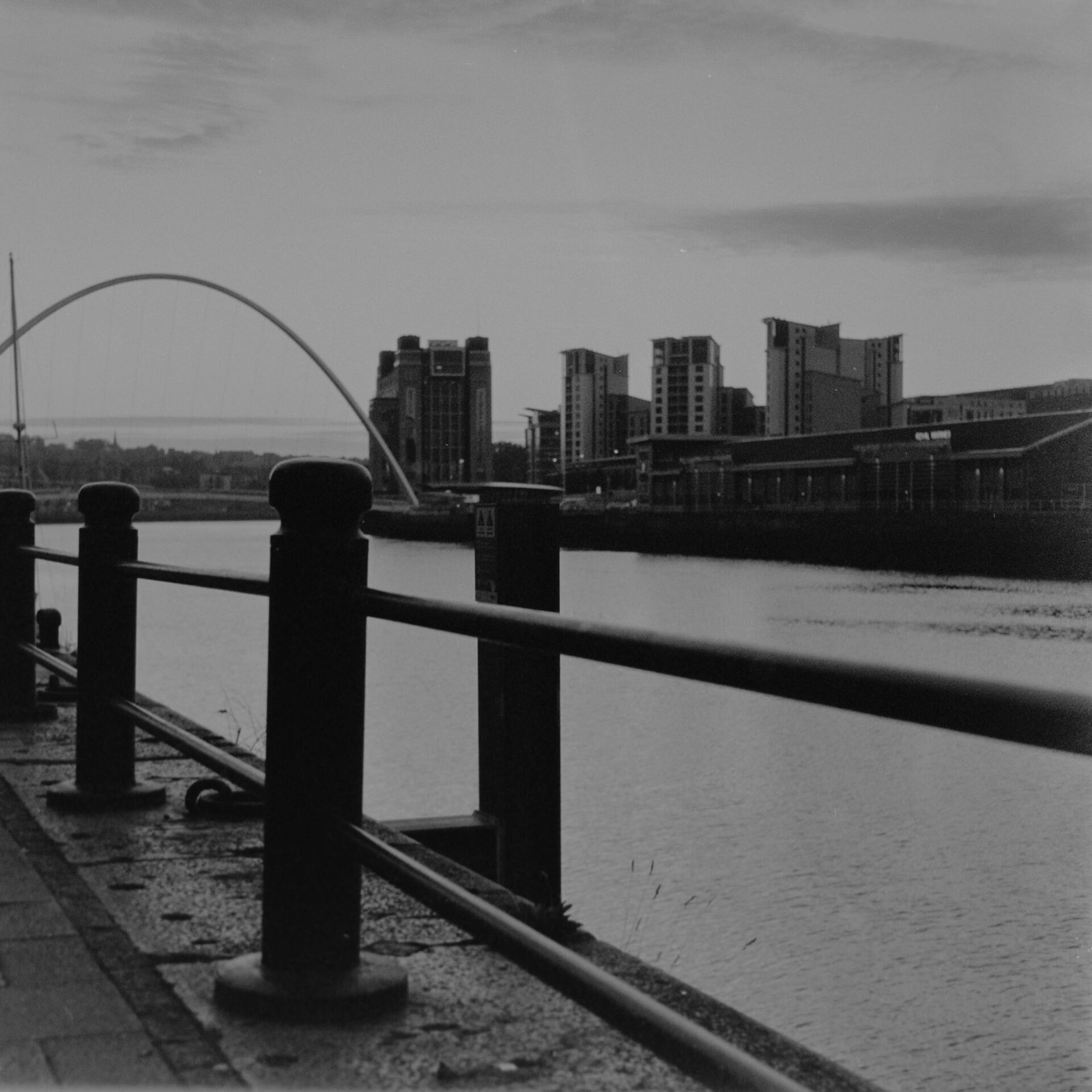 Black and white film photograph of the Gateshead Millennium Bridge and Baltic Centre across the River Tyne at dawn, with quayside railings and bollards in the foreground