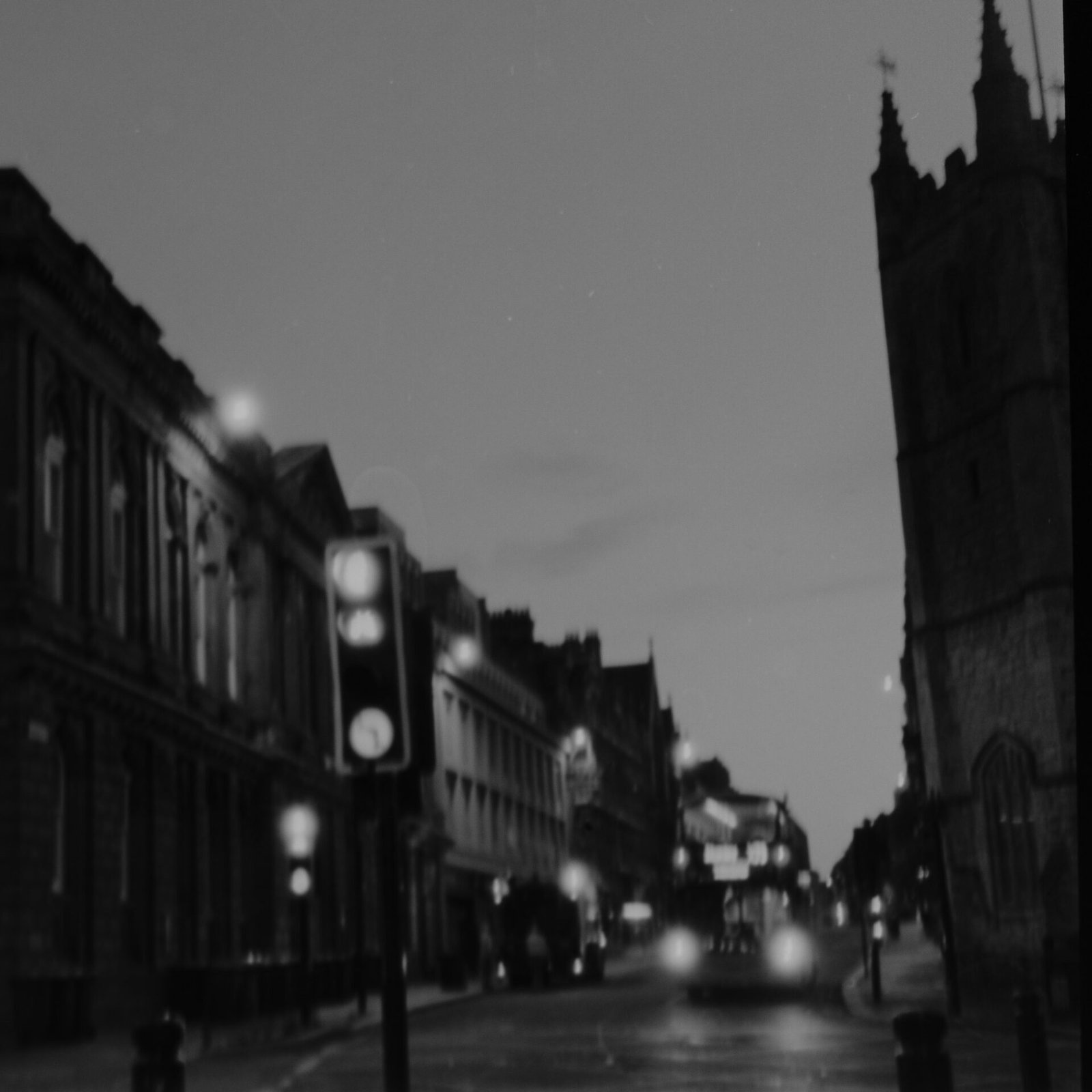 Soft-focus black and white film photograph of a Newcastle city centre street at dawn, traffic lights and headlights glowing beside a Gothic church tower