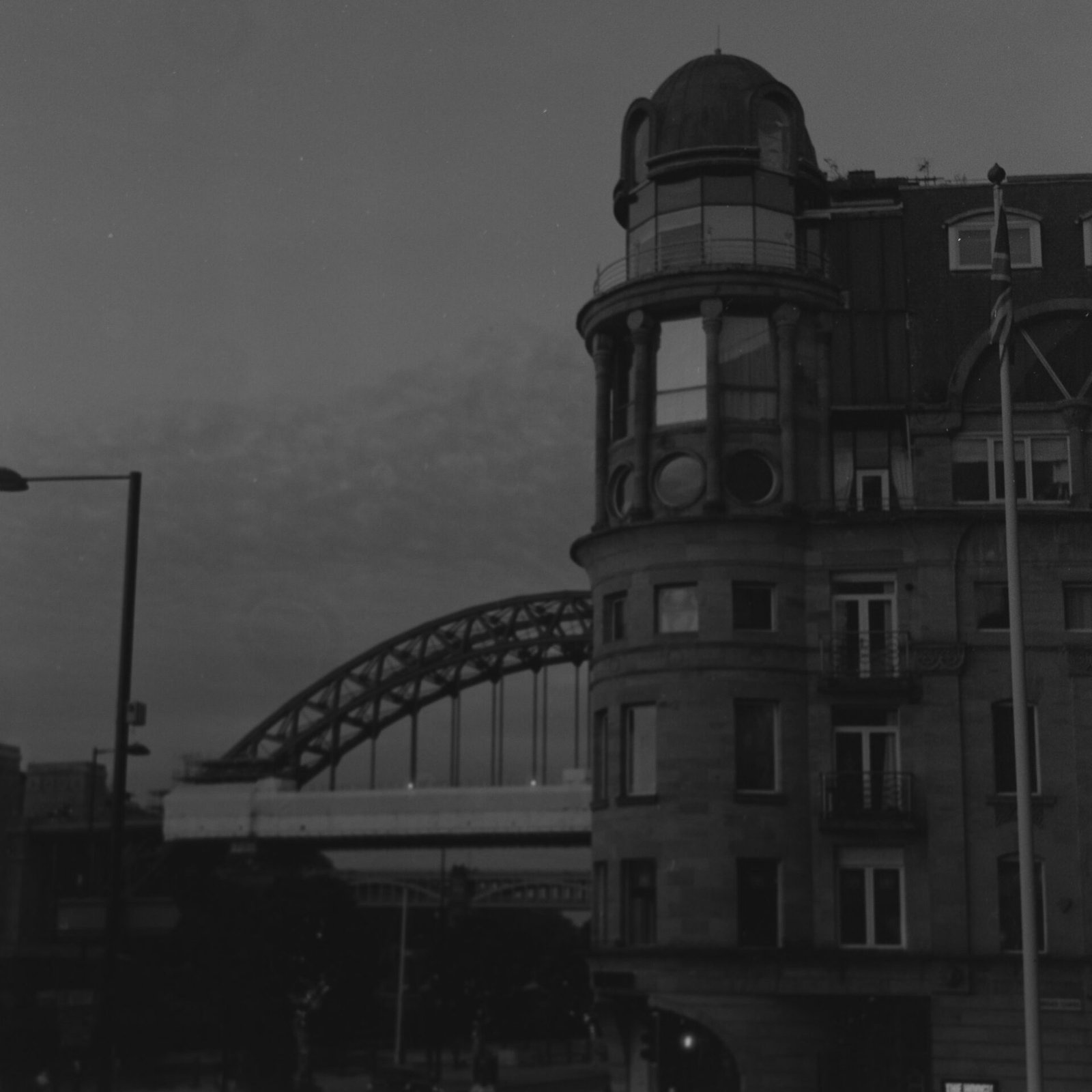 Black and white medium format photograph of an Edwardian building with a domed corner turret on Newcastle’s Quayside, the Tyne Bridge wrapped in scaffolding behind it